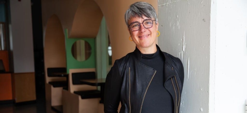 Erin Millar, CEO of Indiegraf, smiling and leaning casually against a white pillar in a modern, warmly lit workspace. She’s wearing a black jacket, round glasses, and yellow earrings.